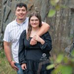 Newly engaged young couple posing out side near a rock wall with greenery in the foreground. Black Hills Photographer Laurel Danley