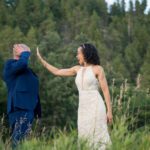 Bride and groom having fun with each other with high 5's with the green pine trees and tall grass around them. Black Hills Photographer Laurel Danley