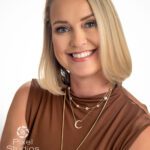 Headshot of a blonde woman with a great big smile in a sleeveless brown blouse and three gold necklaces.