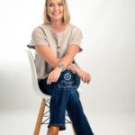 Branding image of a blonde woman sitting in a white chair with writs crossed over her knee on a white backdrop in the studio.