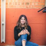 Senior girl sitting up against an orange wall in down urban area with the downspout in the image for a edgy urban look.